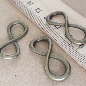 Puede incluir: Tres colgantes con símbolos de infinito de color bronce. Los colgantes son de metal y tienen una superficie ligeramente texturizada. Los símbolos de infinito están dispuestos sobre una superficie marrón claro junto a una regla.