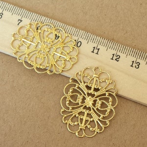 May include: Two gold-colored filigree oval embellishments with intricate, openwork designs. The embellishments are positioned near a wooden ruler marked in inches, suggesting a craft or jewelry-making context. The background is a neutral tan color.