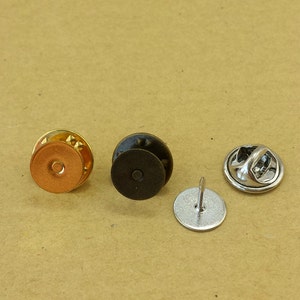 May include: Close-up of four metal pin backs in various colors. There is a gold-colored pin back, a black pin back, a silver pin back with a pin, and a silver pin back with a clasp. The pin backs are on a light brown surface.