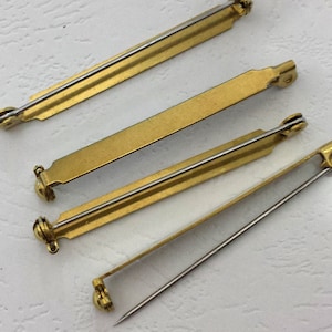 Puede incluir: Cuatro broches dorados con agujas plateadas. Los broches tienen forma rectangular con extremos redondeados y un pequeño cierre en forma de bola. Los broches están dispuestos en diagonal sobre una superficie blanca.