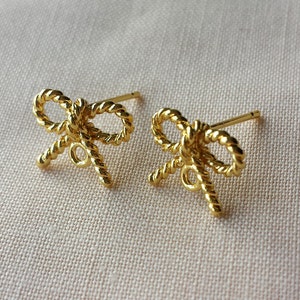 May include: A pair of gold-colored bow-shaped stud earrings. The earrings have a twisted rope design, with a post and backing for secure wear. The bows are detailed and symmetrical, set against a neutral background.