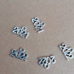 May include: Silver metal charms in the shape of the numbers 2021 and 2022. The charms are scattered on a brown surface. These charms are likely used for jewellery making or other craft projects.