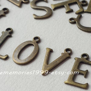 May include: Close-up of bronze metal letter charms spelling "LOVE". The letters have a loop at the top for attaching to jewellery or crafts. Other bronze letters are scattered in the background. The Etsy shop name is visible at the bottom.