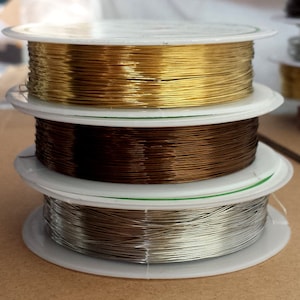 Può includere: Una pila di tre bobine di filo metallico color oro, marrone e argento. Le bobine sono impilate l'una sull'altra, con la bobina dorata in cima. Il filo è avvolto strettamente su bobine di plastica bianca, adatto per la creazione di gioielli o artigianato.