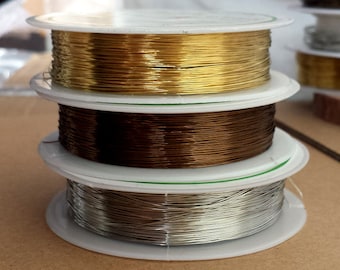 May include: A stack of three spools of wire in gold, brown, and silver. The spools are white with green and black text. The wire is tightly wound and appears to be for jewelry making or crafts.