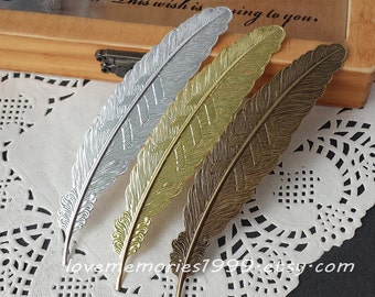 May include: Four metal feather bookmarks in silver, gold, and bronze tones. Each bookmark has a detailed feather design with a textured pattern. The bookmarks are arranged on a white lace doily.