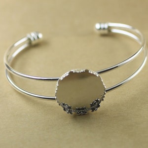 May include: A silver-toned bracelet with a double-band design. The bracelet features a central, round setting with a decorative, scalloped edge, perfect for adding a custom embellishment. The open-ended design allows for easy wear.