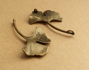 May include: Two antique brass-colored Ginkgo leaf charms. Each charm features a detailed leaf design with a curved stem and a small loop for attaching to jewelry or other crafts. The charms are on a neutral background.