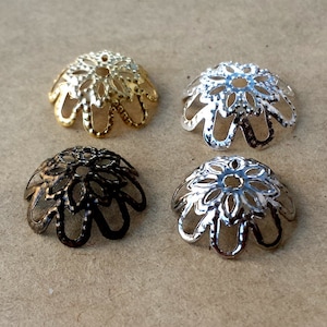 May include: Four filigree bead caps in gold, silver, bronze, and gunmetal. Each cap has a floral design with openwork petals and a small hole at the top. These decorative caps are used in jewelry making.