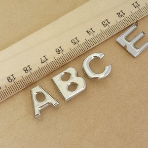 Puede incluir: Letras metálicas plateadas A, B, C y E dispuestas sobre una superficie marrón claro. Una regla de madera con marcas negras se coloca en diagonal, proporcionando una referencia visual.
