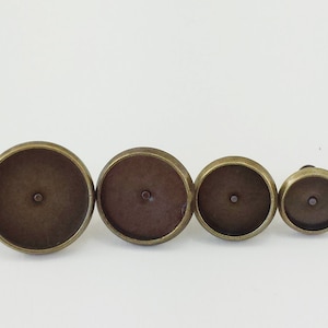 May include: Four antique bronze-colored bezel settings in varying sizes. Each setting has a shallow, circular dish with a small central hole, ideal for crafting. The settings are arranged in a row against a white background.
