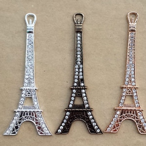 Può includere: Tre ciondoli a forma di Torre Eiffel in argento, bronzo e oro rosa. Ogni ciondolo è decorato con strass trasparenti, che aggiungono brillantezza all'iconico design del monumento parigino. I ciondoli sono esposti su uno sfondo neutro.