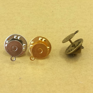 May include: Three metal pin backs in silver, gold, and bronze. Each pin back has a round base with a loop, a central post, and a small disc. The silver and gold pins have decorative star-shaped accents around the edge.