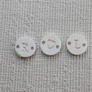 Può includere: Tre piccoli ciondoli circolari color argento. Un ciondolo ha la lettera "R" stampata, un altro ha una forma di cuore e il terzo la lettera "L". Ogni ciondolo ha due piccoli fori su entrambi i lati.