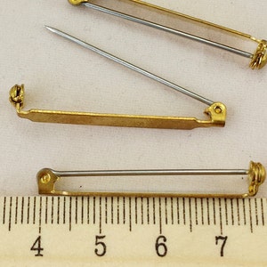 May include: Close-up of several gold-coloured brooch pin backs with silver-coloured needles. The pin backs are rectangular with a rounded end and a clasp. A ruler with inch measurements is in the foreground.