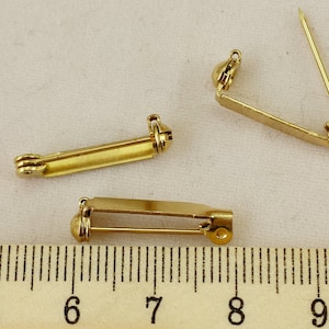 Può includere: Tre retro di spilla color oro sono esposti su uno sfondo chiaro. I retro delle spille sono rettangolari con un meccanismo a spillo e chiusura. Un righello è visibile in primo piano, con misure in centimetri.