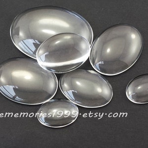 May include: A collection of clear, oval-shaped glass cabochons of varying sizes. The cabochons are transparent, reflecting light and the surrounding environment. The Etsy shop name "lovememories1999.etsy.com" is printed in white text.