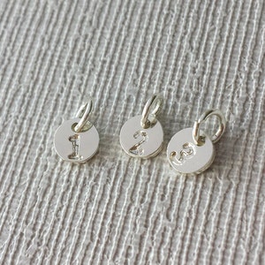 May include: Three small, silver-colored round charms with numbers 1, 2, and 3 engraved on them. Each charm has a small loop at the top for attaching to a chain or bracelet. The charms are displayed on a textured, light gray surface.