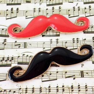 May include: Two mustache-shaped brooches. One is bright red, the other is black. Both have a gold-colored trim. The brooches are set against a background of vintage sheet music.