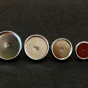 May include: Four round, silver-toned bezel settings of varying sizes. The settings have a flat, circular base with a small hole in the center, designed for crafting or jewelry making. The smallest setting has a dark red center.