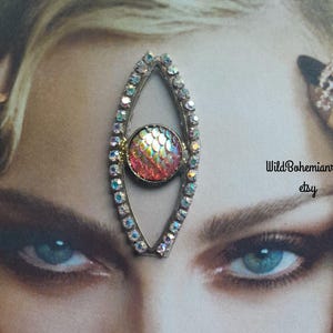 May include: An eye-shaped headpiece featuring a silver-toned frame encrusted with small clear rhinestones and a central circular charm with a pink and orange iridescent scale pattern. The text WildBohemianrose etsy and Always purchase where you see my name on photo are visible.