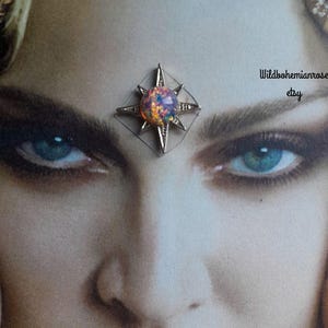 May include: A silver-toned star-shaped bindi or facial ornament featuring a central iridescent opal-like stone with flashes of orange, pink, and blue. The piece is displayed on a printed image of a face with the text Wildbohemianrose etsy visible.