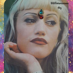 May include: A decorative bindi featuring a teardrop-shaped blue gemstone set in gold-tone metal above a smaller round red gemstone. The image includes the text WildBohemianrose etsy and Always purchase where you see my name on photo.