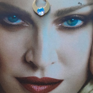 May include: A gold-toned crescent moon bindi featuring a central iridescent crystal. The accessory is displayed on a printed image of a face with blue eyes and red lips. Text on the image reads Wildbohemianrose etsy and Always purchase where you see my name on photo.