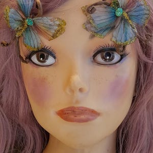 May include: A mannequin head with light purple hair features two decorative butterfly hair clips. The butterflies have blue and pink translucent wings with gold glitter edges and a small green center stone. Text reads: WildBohemianrose etsy.