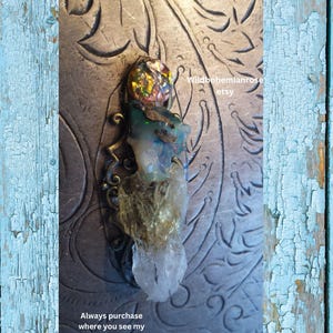 May include: A pendant featuring a clear quartz crystal, a green opal, and a multi-coloured iridescent glass bead. The pendant is attached to a bronze-coloured metal bail. The pendant is displayed on a textured metallic surface. "Wildbohemianrose etsy" is written in white text on the surface.