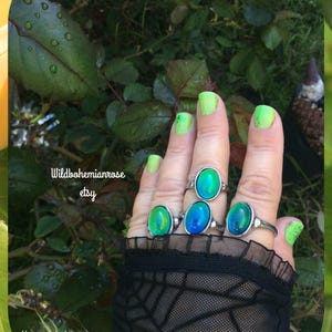 May include: A hand displays four silver rings with oval-shaped, color-changing stones in shades of green and blue. The nails are painted neon green. The image includes the text "Wildbohemianrose etsy" and "Always purchase where you see my name on photo."