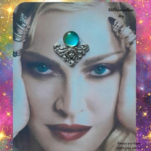 May include: A silver-toned metal hair accessory featuring a central circular teal cabochon stone set above a detailed relief of a face with flowing hair. The accessory is displayed on a printed image of a face with the text WildBohemianRose etsy.