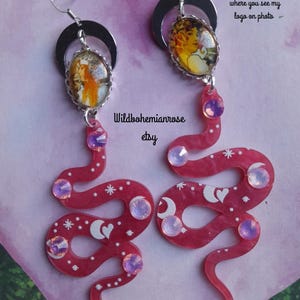 Peut inclure: Une paire de boucles d'oreilles pendantes avec un croissant de lune argenté au-dessus d'un cabochon ovale portant un portrait, relié à un pendentif en forme de serpent rose. Le serpent est orné d'étoiles, de lunes, de cœurs et de cristaux irisés.