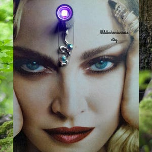 May include: A woman's face with a silver snake-shaped accessory on her forehead, adorned with blue-green gemstones. The text "Wildbohemianrose etsy" is visible. The image has a green and brown background. The accessory has a small purple light.
