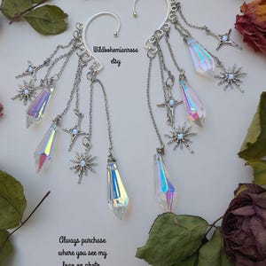May include: A pair of silver-toned ear cuffs featuring dangling chains, iridescent crystal points, and star-shaped charms. The text Wildbohemianrose etsy and Always purchase where you see my logo on photo is visible on the white background.