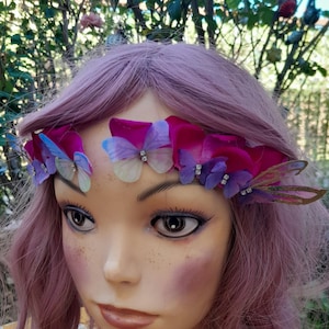 May include: A headpiece featuring vibrant magenta fabric petals interspersed with small, translucent butterflies in shades of blue, purple, and white, accented with clear rhinestones, worn on a mannequin with light pink hair.