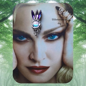May include: An elaborate silver hair clip with a central iridescent stone and purple and blue embellishments. The clip has a dripping design and is set against a portrait with blue eyes and red lips. The Etsy shop name "Wildbohemianrose" is visible.
