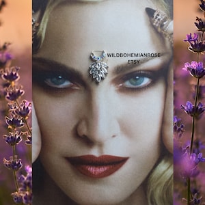 May include: A silver and crystal forehead chain with a teardrop design. The chain is adorned with clear rhinestones and blue accents. The chain is worn by a woman with blonde hair and blue eyes. The image is set against a backdrop of lavender flowers.