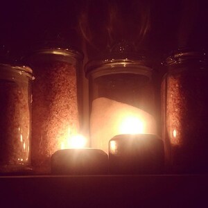 May include: Four glass jars filled with different types of salt, illuminated by candlelight. The jars are arranged in a row on a shelf.