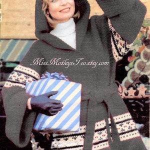 May include: A woman wearing a gray hooded crocheted sweater with a white turtleneck underneath. The sweater has a white and brown geometric pattern on the sleeves and hem. She is holding a blue and white striped gift box and wearing black gloves. The text "Miss Mortleys Too.etsy.com" is visible on the sweater.