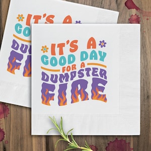 Op de afbeelding: Twee witte papieren servetten met een kleurrijk grafisch ontwerp. De tekst op de servetten luidt "It's a good day for a dumpster fire".
