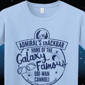 A light blue Star Wars t-shirt with a navy blue graphic print of a logo for a snack bar called 'Admiral's Snack Bar.' The logo features Admiral Ackbar, with the text 'Home of the Galaxy Famous Obi-Wan Cannoli.' The text 'Served 'Vader-style' (sliced in half)' is printed below the logo.