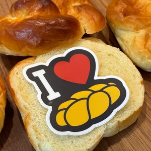 May include: An assortment of baked goods, including golden-brown challah bread and sliced bread. A sticker on the bread reads "I ❤️ Challah" with a red heart and yellow challah illustration. The bread is on a wooden surface.