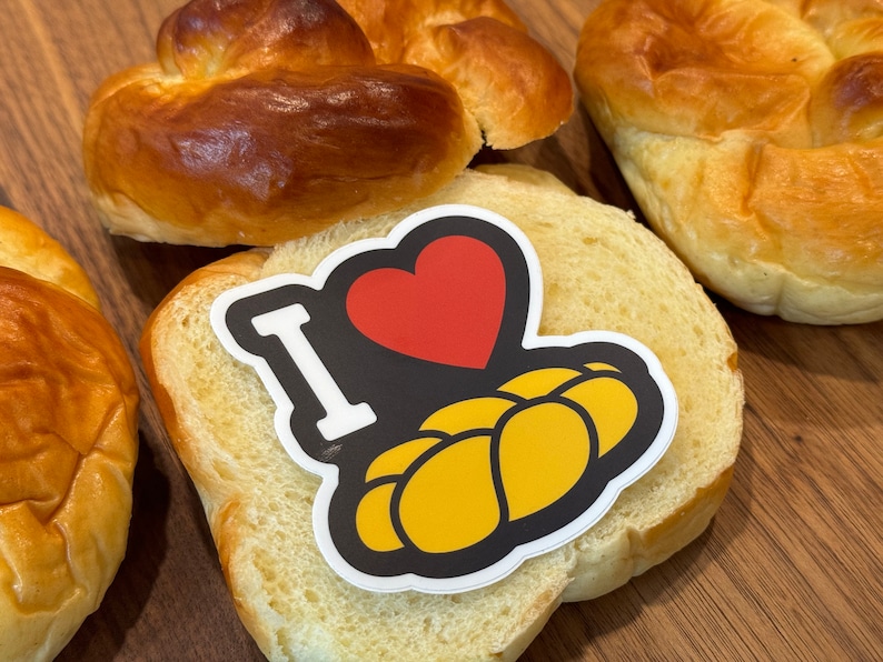 May include: An assortment of baked goods, including golden-brown challah bread and sliced bread. A sticker on the bread reads "I ❤️ Challah" with a red heart and yellow challah illustration. The bread is on a wooden surface.