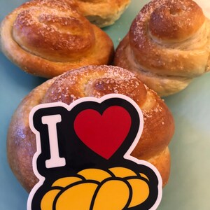 May include: Four golden-brown, spiral-shaped baked goods, sprinkled with coarse sugar, rest on a light blue surface. One bun has a sticker that says "I" with a red heart and a yellow bread graphic.