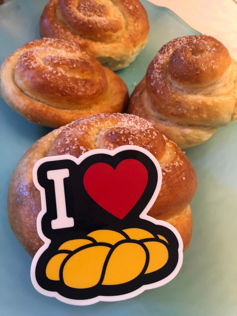 May include: Four golden-brown, spiral-shaped baked goods, sprinkled with coarse sugar, rest on a light blue surface. One bun has a sticker that says "I" with a red heart and a yellow bread graphic.
