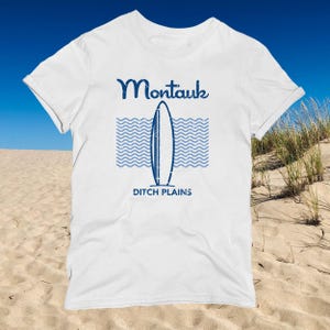 Puede incluir: Camiseta blanca con un gráfico azul que representa una tabla de surf, olas y el texto "Montauk" encima de "Ditch Plains". El diseño tiene un aspecto envejecido y vintage. La camiseta está hecha de un material suave y ligero.
