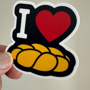 May include: A sticker with a black background, a red heart, and the white letter "I". Below the heart is a yellow, braided loaf of bread. The sticker has a white border.
