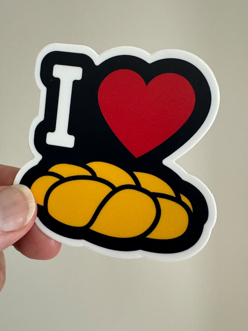 May include: A sticker with a black background, a red heart, and the white letter "I". Below the heart is a yellow, braided loaf of bread. The sticker has a white border.