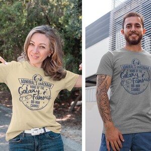 A woman and man wearing a Star Wars t-shirt with a navy blue graphic print of a logo for a snack bar called 'Admiral's Snack Bar.' The logo features Admiral Ackbar, with the text 'Home of the Galaxy Famous Obi-Wan Cannoli.' The text 'Served 'Vader-style' (sliced in half)' is printed below the logo.